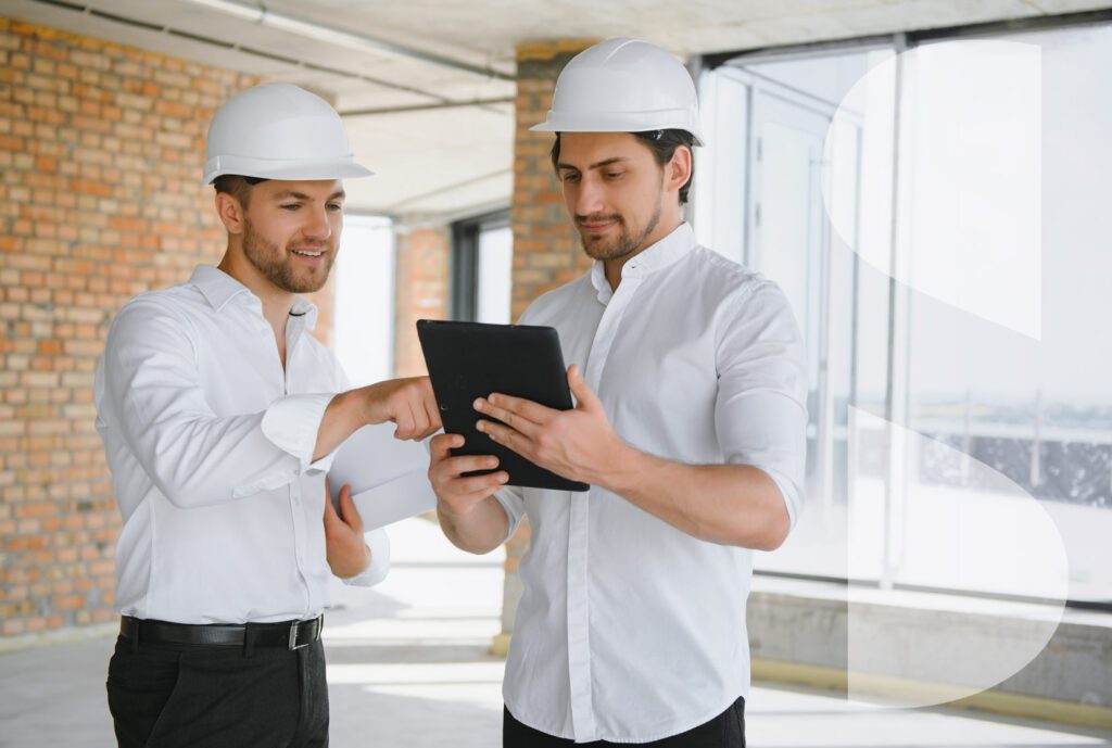 Two architects in construction hats conduct business on a tablet at a construction site.